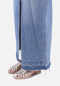Falda maxi de mezclilla con dobladillo deshilachado y abertura lateral, combinada con sandalias metálicas trenzadas que tienen un bajo tacón de madera y dedos al aire.