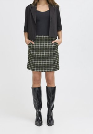 Nainen, jolla on musta lyhyt bleiseri, musta toppi, vihreä-valkoruudullinen taskullinen hame ja polven korkuiset mustat nahkasaappaat, seisomassa valkoisella taustalla.