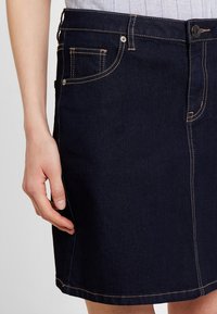 Džínová sukně v tmavě modré barvě se střihem rovného střihu. Obsahuje zadní kapsu, viditelné švy v světle hnědé barvě a zapínání na knoflík v pase.