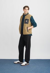 Tan fleeceväst med svarta detaljer och bröstficka, buren över en mörkgrön skjorta, ihop med svarta byxor och vita sneakers på ett blått golv.