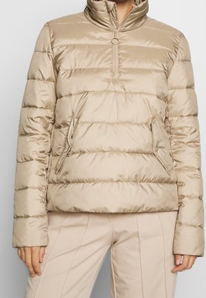 Beige gewatteerd jack met horizontale doorgestikte lijnen, halve rits aan de voorkant met ronde ritskop, zijzakken, gedragen met een beige broek.
