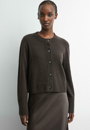 Woman wearing a dark brown buttoned cardigan with long sleeves and a matching brown skirt against a plain light background.