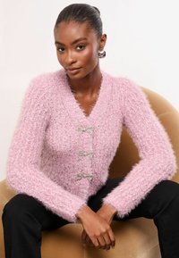Rozā pūkains kardigans ar V-veida apkakli un trim dekoratīvām sudraba loku aizdarnēm. Ar garām piedurknēm un mīkstu, teksturētu audumu. Valkāts virs melnajām biksēm.
