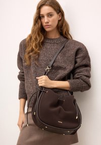 Brun tygaxelväska med en mjuk, rundad form, detaljer i guld och dragsnörning. Modellen håller den mot en beige outfit.