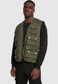 Oliwkowa kamizelka robocza z wieloma przednimi kieszeniami, srebrnymi napami, kieszenią na zamek oraz metalowym detalem w postaci pierścienia. Noszona na czarnej koszuli.