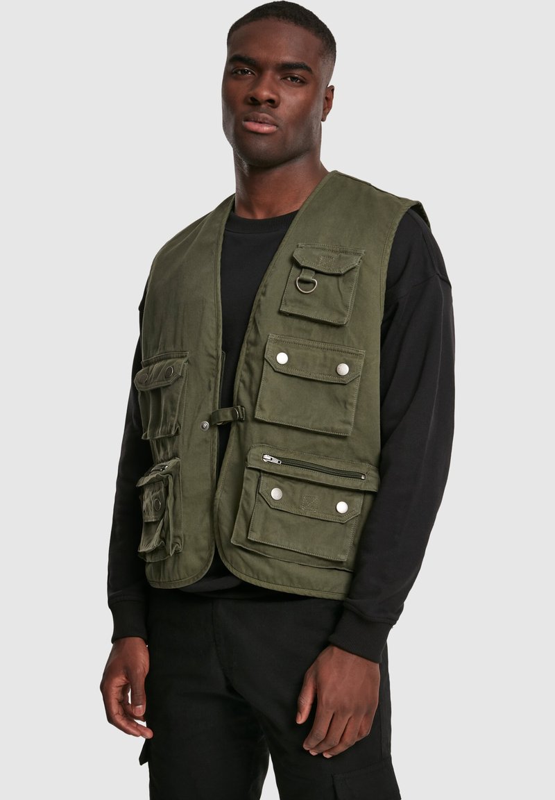 Oliwkowa kamizelka robocza z wieloma przednimi kieszeniami, srebrnymi napami, kieszenią na zamek oraz metalowym detalem w postaci pierścienia. Noszona na czarnej koszuli.