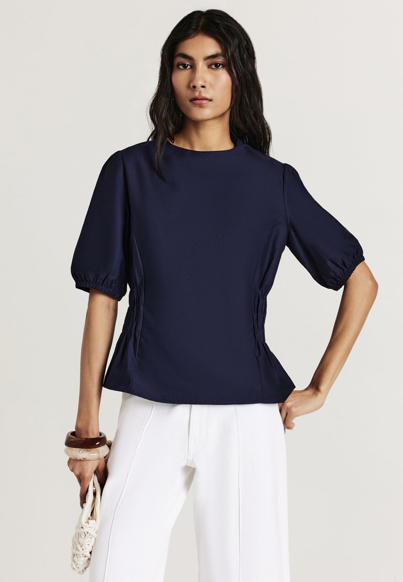 Blusa blu navy con scollo rotondo, maniche a sbuffo e arricciature laterali. Realizzata in un tessuto liscio, abbinata a pantaloni bianchi a gamba larga.