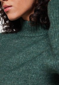 Femme aux cheveux bouclés et foncés portant un pull vert en laine à col roulé, lèvres légèrement entrouvertes, tournée vers la gauche avec un bras levé près de la tête.