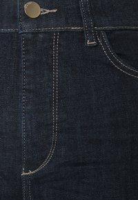 Jeans de mezclilla oscura con costuras en contraste, un bolsillo frontal y cierre con botón de metal. Diseño estándar de cinco bolsillos.