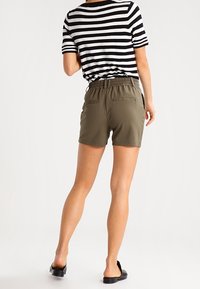 Chemise rayée noire et blanche à manches courtes, associée à un short vert olive avec taille élastique et poches arrière. Chaussures plates noires.