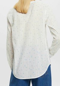 Persona vestida con una blusa blanca de manga larga con pequeños estampados florales azules y jeans azules, vista desde atrás.