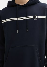 Sudadera azul marino con rayas horizontales blancas, cordones y bolsillo frontal. Presenta un logotipo con "EST. 2007" debajo. Tejido suave, diseño casual.