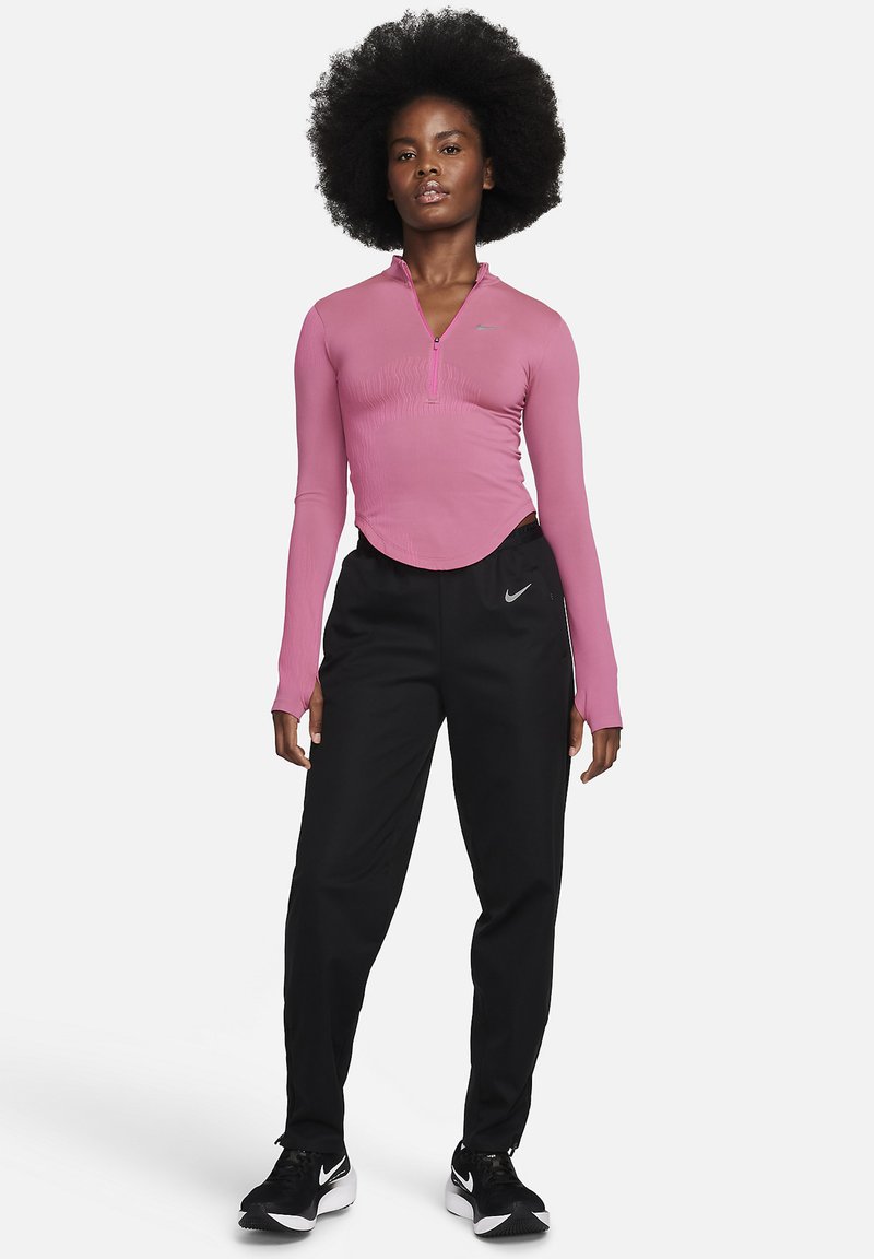 Mujer con un peinado afro natural, vistiendo una camiseta rosa de manga larga con cremallera, pantalones de chándal negros de Nike y zapatillas negras de Nike, se encuentra de pie de frente.