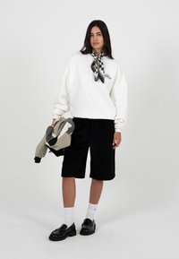 Femme portant un sweat-shirt blanc, un short noir mi-genou, une écharpe à carreaux, des chaussettes blanches et des mocassins noirs, tenant une veste color-block.