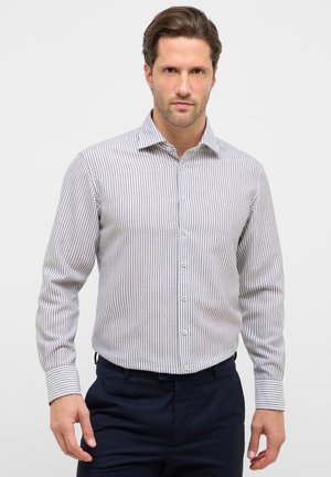 Uomo che indossa una camicia a maniche lunghe a righe verticali bianche e grigie e pantaloni blu scuro su uno sfondo bianco semplice.
