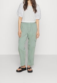 Světlomyslné šedé tričko s nadýchanými rukávy z krajky, zkombinované s volnými krojovými kalhotami v pastelově zelené barvě, které mají boční kapsy a elastický pas. Černé žabky.