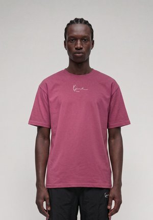 Homme aux cheveux tressés portant un t-shirt rose à manches courtes et un pantalon noir, debout devant un fond clair uni.