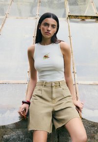 Canottiera bianca a costine con motivo ricamato verde e rosso, abbinata a pantaloni corti beige chiaro a vita alta con orlo sfrangiato.