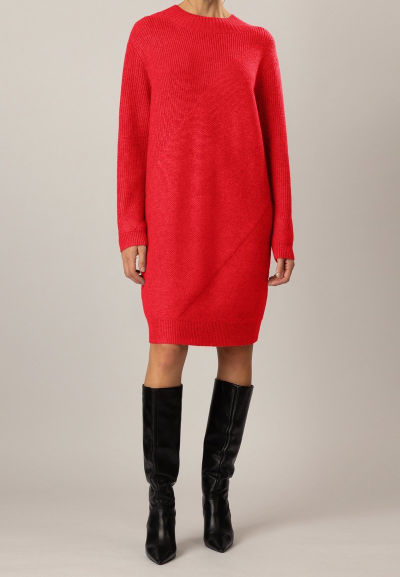 Femme portant une robe en maille rouge à manches longues et des bottes en cuir noires montant jusqu'aux genoux, debout devant un fond uni.