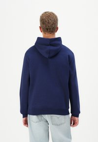 Persona di spalle che indossa un semplice hoodie blu navy e jeans chiari su uno sfondo bianco uniforme.