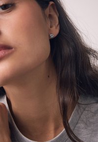 Close-up of a woman wearing a silver twisted knot stud earring with long dark hair and natural skin tone against a light background.
