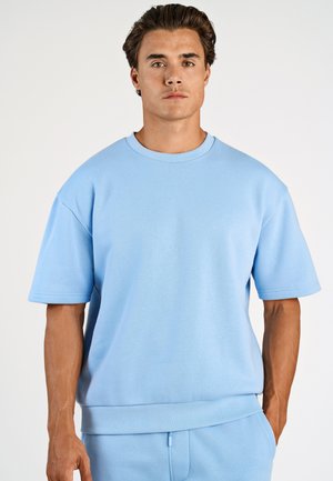 Man met kort bruin haar, gekleed in een lichtblauw sweatshirt met korte mouwen en bijpassende broek, staand tegen een effen witte achtergrond.