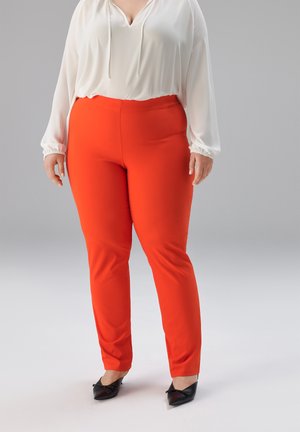 Frau trägt leuchtend orangefarbene, eng anliegende Hose, eine langärmelige weiße Bluse und schwarze spitze Schuhe, steht vor einem grauen Hintergrund.