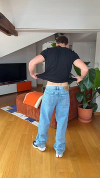 Mężczyzna w czarnej koszuli i dżinsach podnoszący koszulę w salonie z brązową kanapą, rośliną, telewizorem i drewnianą podłogą.