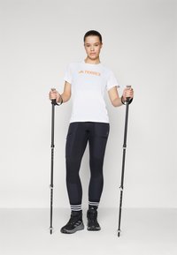 Hvit TERREX t-skjorte med oransje logo, svarte tettsittende leggings med teksturerte paneler, og svarte tursko. To turstaver holdt i hendene.