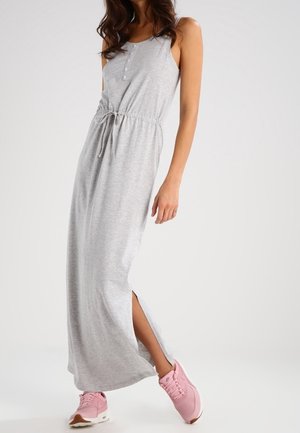 Femme portant une robe maxi grise sans manches avec une fente latérale et des baskets roses, debout devant un fond clair uni.