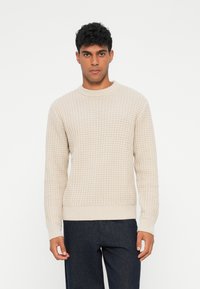 Maglione beige a maglia con motivo testurizzato, scollatura rotonda e polsini e orlo a coste, abbinato a pantaloni scuri.