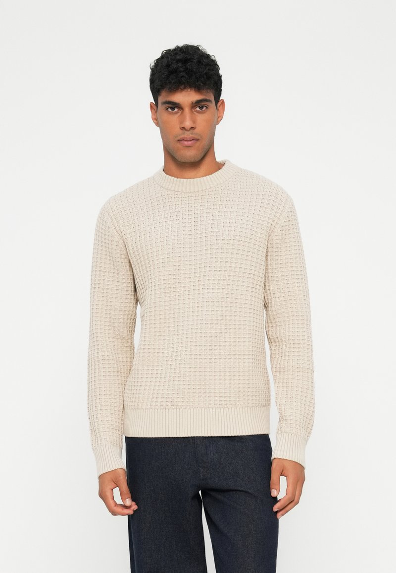 Maglione beige a maglia con motivo testurizzato, scollatura rotonda e polsini e orlo a coste, abbinato a pantaloni scuri.