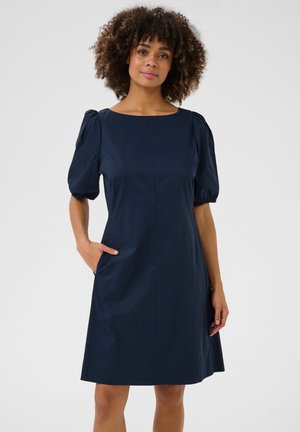 Kobieta o kręconych włosach, ubrana w granatową sukienkę do kolan z bufkami, stoi z jedną ręką w kieszeni na jednolitym tle.