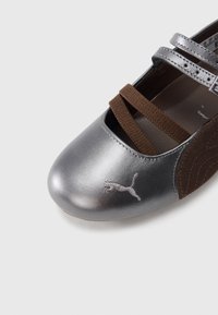 Chaussure argent métallisé avec brides élastiques marron, bout arrondi, accents en suède et détail logo argenté sur le côté. Surfaces lisses et texturées.