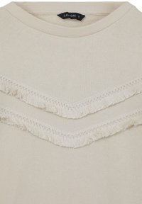 Pull en maille beige à col rond, présentant deux rangées en V de franges décoratives sur la poitrine, étiquette taille small visible.