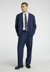 Costume bleu marine avec un revers cranté, fermeture à deux boutons et pantalon plissé. Assorti à une chemise blanche et une cravate rayée. Chaussures noires.