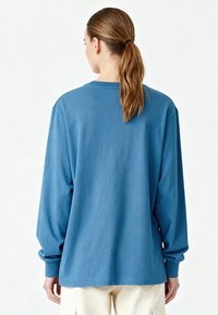 Femme avec une queue de cheval châtain clair portant une chemise bleue à manches longues et un pantalon beige clair, de dos sur un fond blanc uni.