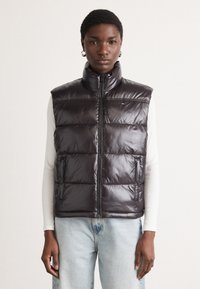 Tamno ljubičasti puffer prsluk s visokim ovratnikom, prednjim zatvaračem, dijagonalnim kvadratnim uzorkom i bočnim džepovima, nošen preko svijetlog dugog rukava.