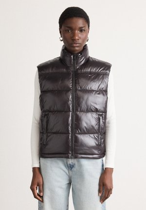 Dark purple puffer vest with high collar, front zipper, diagonal quilted pattern, and side pockets, worn over a light-colored long-sleeve top.