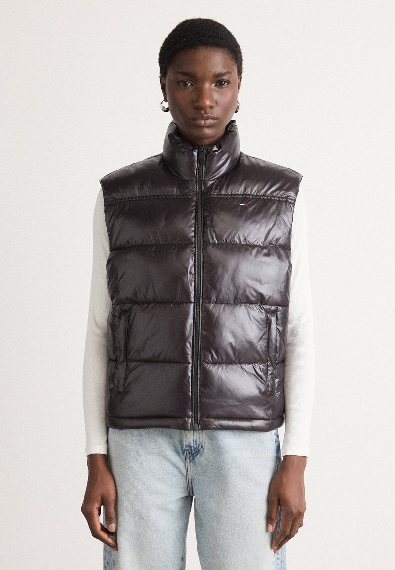 Tamno ljubičasti puffer prsluk s visokim ovratnikom, prednjim zatvaračem, dijagonalnim kvadratnim uzorkom i bočnim džepovima, nošen preko svijetlog dugog rukava.