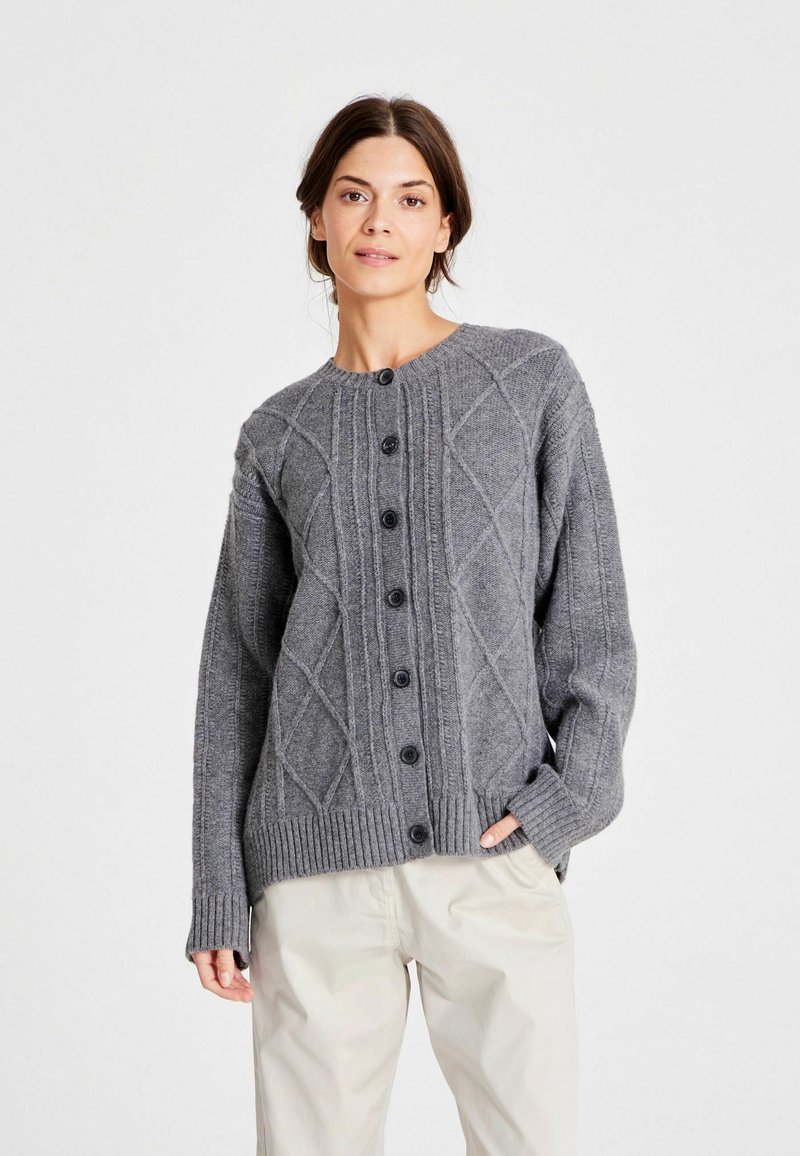 Femeie îmbrăcată cu un cardigan căptușit călcat gri, cu nasturi, și pantaloni bej deschis, stând cu o mână în buzunar pe un fundal simplu.