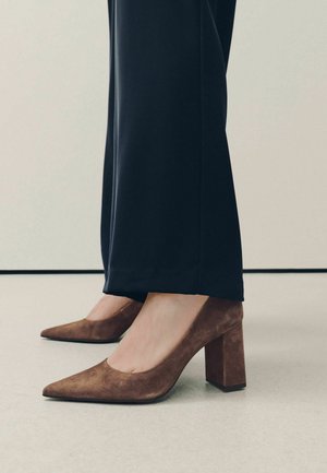 Person wearing brown suede block heel shoes with pointed toes and black wide-leg pants standing on a light floor.