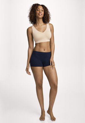 Frau mit lockigem Haar steht barfuß, trägt einen beigen Sport-BH und dunkelblaue Shorts vor einem schlichten hellen Hintergrund.