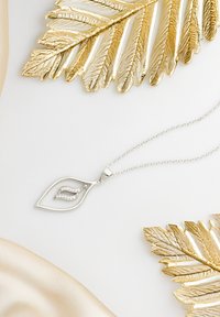 Sølvbladformet vedhæng med tre lodrette linjer af små klare sten, på en fin kæde, sat mod guldblad accenter og blød stof.