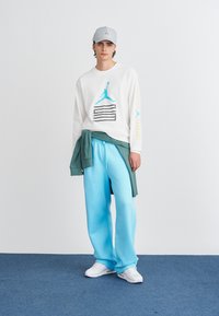 Camicia bianca a maniche lunghe con logo blu, pantaloni della tuta azzurri, maglione verde legato in vita e cappellino grigio. Le sneakers bianche completano il look.