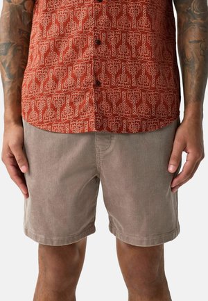 Mann trägt hellbraune Cord-Shorts und ein rotes, gemustertes Hemd mit Knöpfen, steht mit entspannt an den Seiten hängenden Händen.