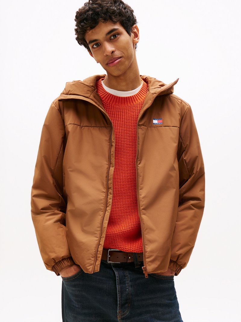 Giacca cappuccio marrone con polsini elasticizzati, indossata sopra un maglione arancione texturizzato. Jeans scuri e una cintura completano l'outfit. Presenta il logo del marchio sul petto.