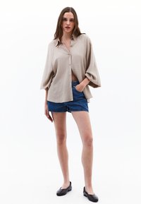 Camicia beige con bottoni e maniche a palloncino, abbinata a pantaloni corti in denim scuro. Il modello indossa delle scarpe nere e ha capelli lunghi e mossi.