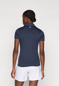 Námořnická modrá polo košile s krátkým límcem, vyrobená z hladkého materiálu, s logem na zadní straně. Vybaveno bílými šortkami.