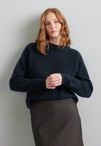 Pull en marine sombre en tissu doux, avec une coupe légèrement ample, col rond et poignets côtelés, associé à une jupe crayon marron.